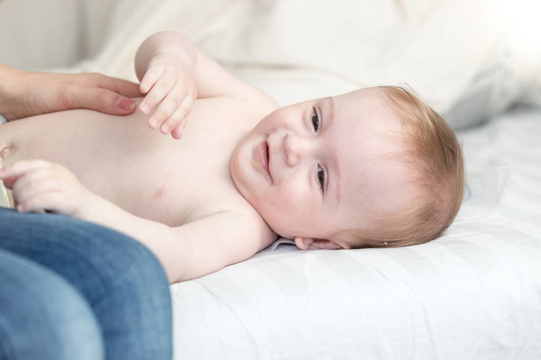 Portrait de mignon bébé garçon souriant allongé sur le lit tandis que la mère le caresse
 - Photo, image