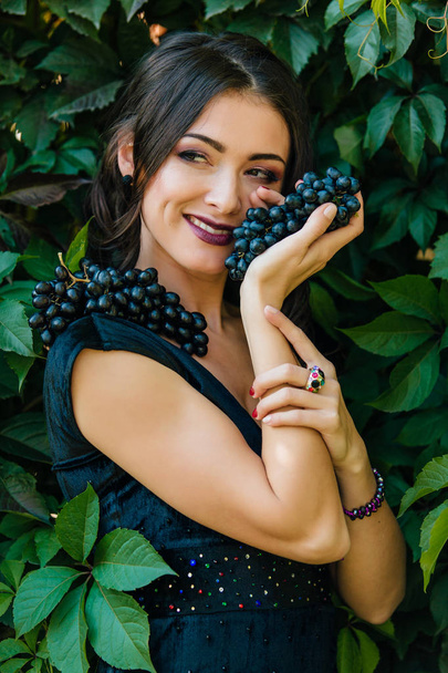 Portrait of young sexy brunette in black dress with black grapes. . - Fotó, kép