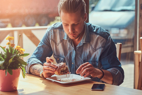 Guapo hipster con un corte de pelo elegante y barba se sienta en una mesa, decidió cenar en un café al lado de la carretera, comiendo una hamburguesa
. - Foto, imagen