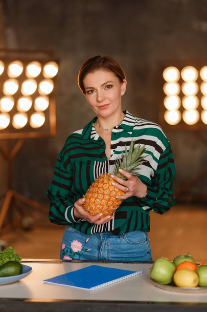 Fitness, home and diet concept. Young woman holding pineapple standing at kitchen table with retro bulb light on background - Foto, imagen