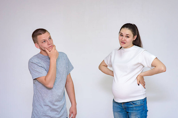 family relationships. Portrait of young married couple: husband and pregnant wife communicate in studio on white background. They stand directly in front of the camera and show different emotions - 写真・画像
