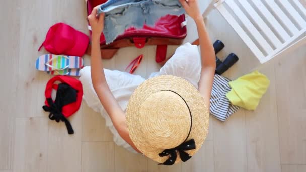 Szőke nő kalapot és fehér ruhát a padlón ül, és csomagolás ruhák utazótáska felülnézet - Felvétel, videó