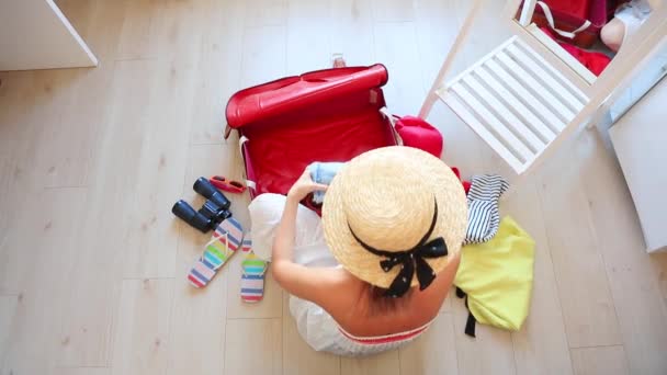 Κάτοψη του ξανθιά γυναίκα με καπέλο και λευκό φόρεμα κάθεται στο πάτωμα και συσκευασία ρούχων σε τσάντα ταξιδιού - Πλάνα, βίντεο