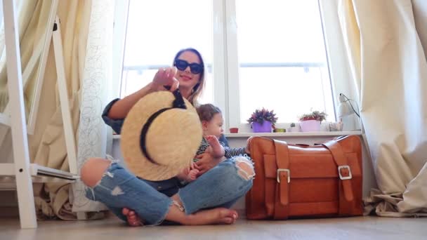 Mujer sentada en el suelo con su hijo cerca de la bolsa de viaje y tomando sombrero
 - Metraje, vídeo