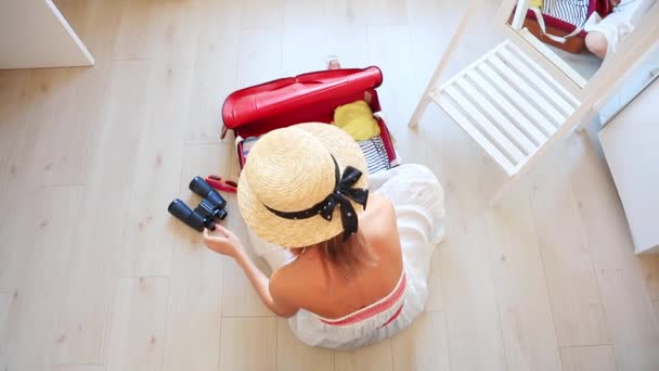 Szőke nő kalapot és fehér ruhát a padlón ül, és csomagolás ruhák utazótáska felülnézet - Felvétel, videó