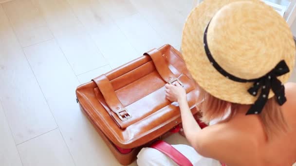 Pohled shora na blond žena v klobouku a bílé šaty sedí na podlaze a balení oblečení do cestovní tašky - Záběry, video
