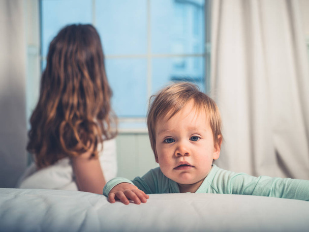 A cute little boy is looking at the camera with his mother in the background in bed by the window - Foto, afbeelding