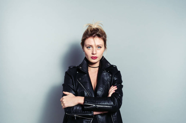 Close up portrait of a pretty blonde woman, wearing black biker jacket, looking at camera, against plain studio background - Photo, image