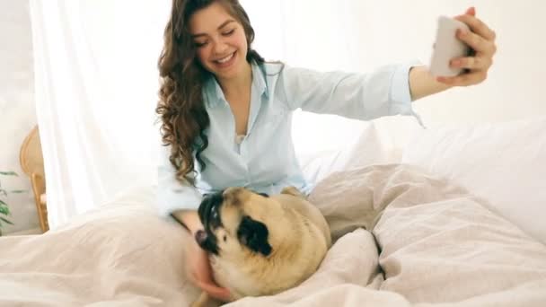 attractive brunette woman in bed taking selfie with her small dog pug. Video footage. Good weekend morning mood. Playing with funny pet - Video