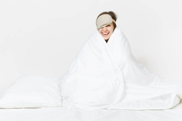 Zábava unavený mladá žena sedí v posteli s spánku masku, list, polštář, zabalení do deky izolovaných na bílém pozadí. Krása žena tráví čas v pokoji. Odpočinek, klid, dobrá nálada koncept. Kopírovat prostor - Fotografie, Obrázek