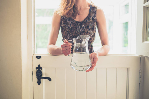 A young woman is standing by a stable door with a jug of water on a sunny day - Foto, imagen
