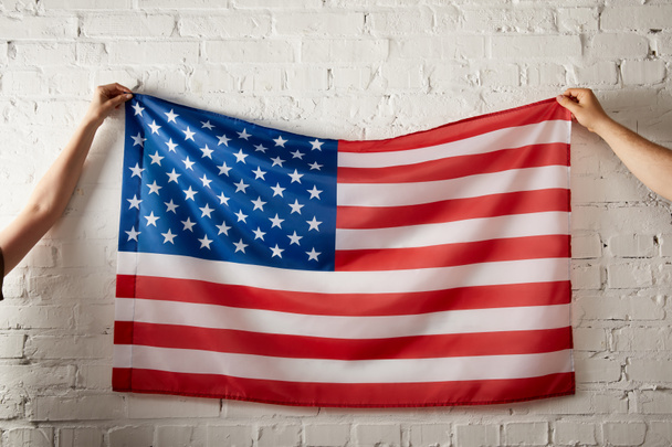 レンガの壁にアメリカの国旗を保持している男女の画像をトリミング  - 写真・画像