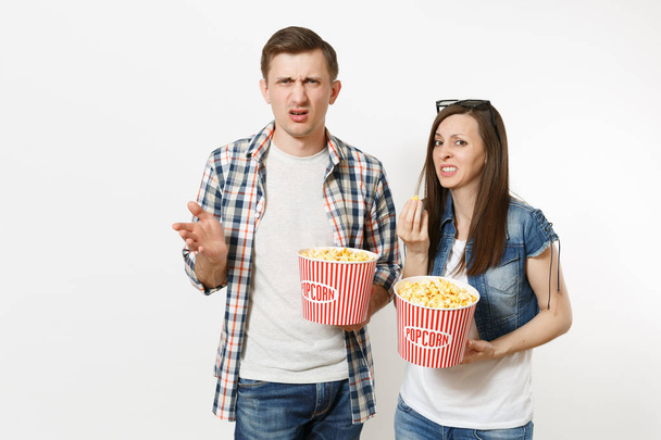 Young irritated dissatisfied couple, woman and man in 3d glasses and casual clothes watching movie film on date, holding buckets of popcorn isolated on white background. Emotions in cinema concept - Photo, Image