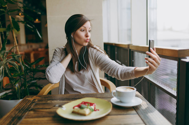 junge Frau sitzt allein im Café am Tisch mit einer Tasse Cappuccino, Kuchen und entspannt sich in der Freizeit im Restaurant. junge Frau beim Selfie mit dem Handy, Ruhe im Café. Lifestylekonzept - Foto, Bild