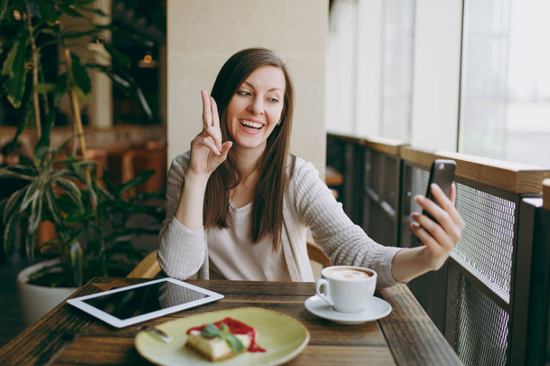 Женщина сидит в кафе с чашкой капучино, торт, делает селфи на мобильный телефон, расслабляется в ресторане в свободное время. Женщина, работающая на планшетном компьютере, отдыхает в кафе. Концепция образа жизни
 - Фото, изображение