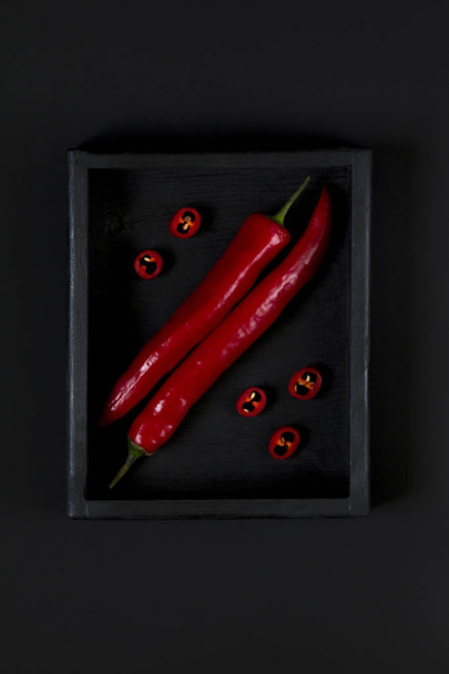 Chili peper in een houten doos op een zwarte achtergrond. Zwart en rood. Abstractie. Bovenaanzicht. - Foto, afbeelding