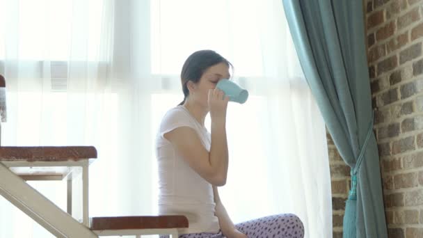 Mladá žena pije kávu na schodišti u okna - Záběry, video