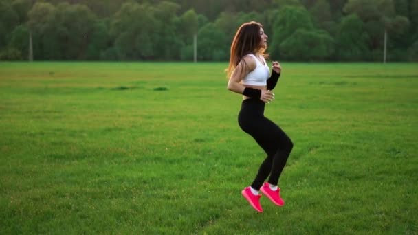 Napnyugtakor a egy mezője a fűben, egy fehér top és rózsaszín cipők, sportos lány teszi a bemelegítés fut csípő feloldása előtt. Sport. közelről. Fű cipők - Felvétel, videó