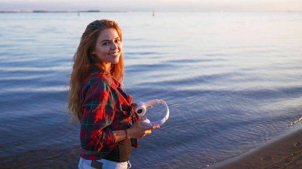 Happy attractive red-haired girl is enjoying favorite music on walk. Beautiful young woman in shirt uses headphones - Φωτογραφία, εικόνα