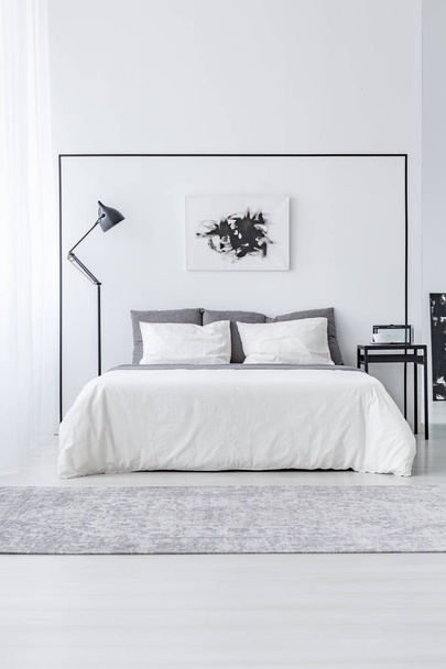 Black and white poster above bed between table and lamp in simple bedroom interior - Fotografie, Obrázek