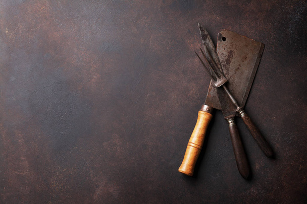 Мясник и кухонная утварь. Винтажный мясной тесак, нож и вилка над каменной доской. Вид сверху с пространством для вашего текста
 - Фото, изображение