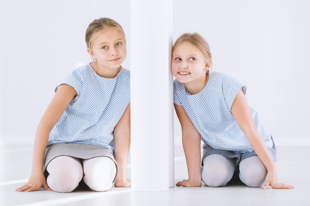 Portrait of young, happy twin sisters kneeling and leaning against opposite sides of a white wall - Foto, Bild