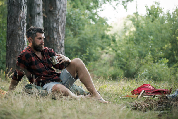 Hipster drink tea or coffee at bonfire. Bearded man relax with mug at campfire. Guy enjoy camping in forest. Summer vacation concept. Camping and hiking and travel. - Фото, изображение