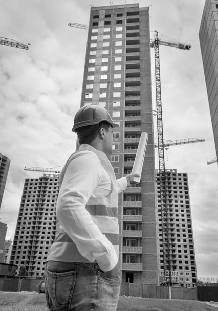 Imagem em preto e branco do arquiteto masculino em pé no canteiro de obras e apontando para o edifício em construção
 - Foto, Imagem