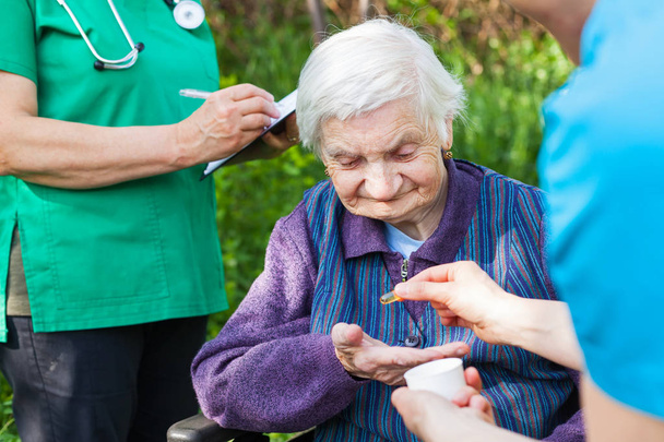 Elderly ill woman receiving pills from nurse outdoor, doctor writing medical prescription in the background - Photo, Image
