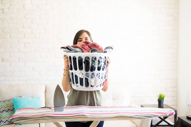 Housewife covering face with heavy laundry basket and keeping above iron table - Photo, Image