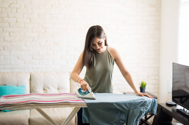 Vrolijke jonge huisvrouw dragen casuals Strijkservice mannelijke shirt op strijkplank - Foto, afbeelding