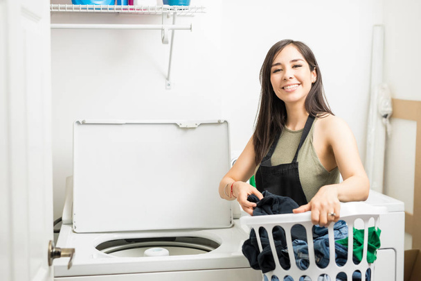 junges Hausmädchen in schwarzer Schürze steckt Kleidung in Waschmaschine - Foto, Bild