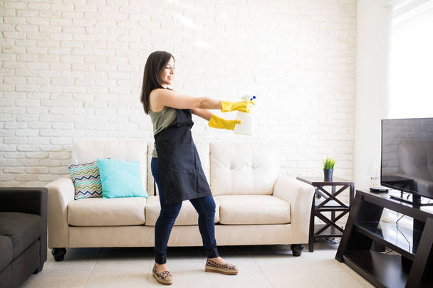 Nuori moderni nainen leikkisä tuulella yllään musta esiliina ja keltainen lateksi käsineet käyttäen spray aseena puhdistaa olohuone
 - Valokuva, kuva