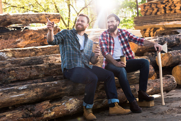 Lächelnder Holzfäller mit Kaffeetasse zeigt mit dem Finger auf Partner mit Axt, der auf Baumstämmen am Sägewerk sitzt  - Foto, Bild