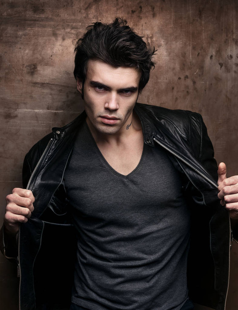 Handsome young man in stylish black leather jacket. Fashion model in dark clothes on a background of old metal wall - Photo, image