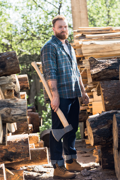 bearded lumberjack  in checkered shirt standing with axe at sawmill  - Fotó, kép