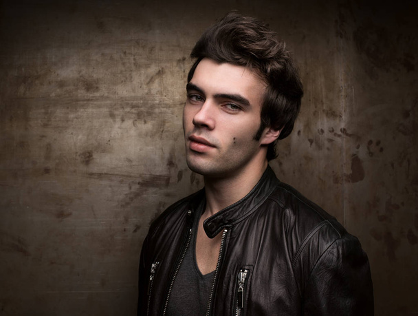 Portrait of a young man against the background of a metal wall with free space. Stylish hairstyle, black leather jacket - Foto, Imagen