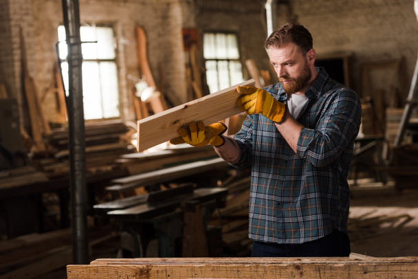 bearded carpenter in protective gloves checking wooden plank at sawmill - Foto, afbeelding