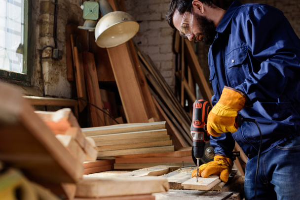 bärtiger Handwerker in Schutzhandschuhen und Schutzbrille bohrt Holzplanke in Tischlerei  - Foto, Bild