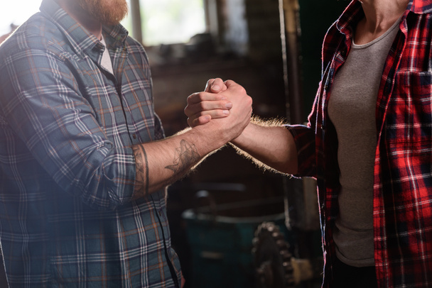 cropped image of carpenter with tattooed hand shaking hand of partner at sawmill  - Photo, Image
