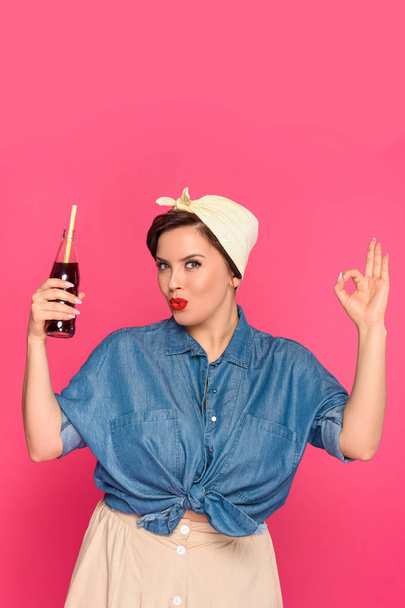 beautiful pin up woman holding glass bottle with drinking straw and showing ok sign isolated on pink - Fotografie, Obrázek