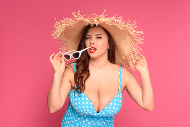 sexy mujer joven en traje de baño y sombrero de paja morder gafas de sol y mirando a la cámara aislada en rosa
  - Foto, Imagen