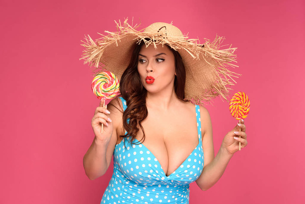doubtful overweight woman in swimsuit holding candies isolated on pink - Фото, зображення