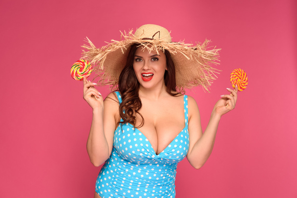 beautiful size plus model in swimsuit holding lollipops and smiling at camera isolated on pink - Photo, Image