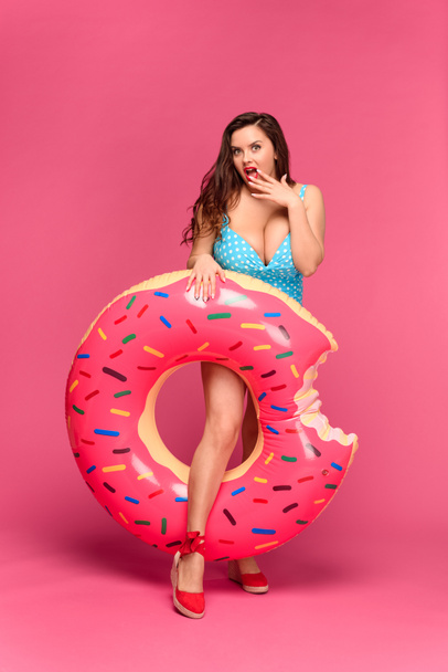 beautiful surprised woman in swimsuit holding swimming ring and looking at camera isolated on pink - 写真・画像