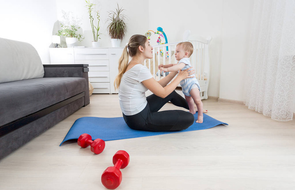 Beautiful young woman doing fitness exercises with her baby boy on floor at home - Zdjęcie, obraz