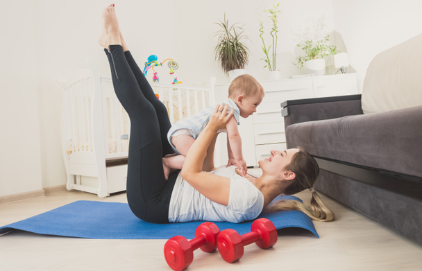 Hermosa mujer joven sosteniendo a su bebé niño y haciendo ejercicio en el suelo en casa
 - Foto, Imagen