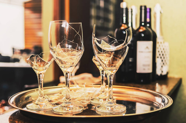 Gebroken wijnglas op een dienblad met sommige wijn en champagne flessen vaag op de achtergrond. Gebroken glas voor een feest of de viering. - Foto, afbeelding