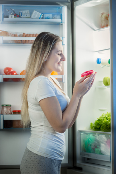 Lächelnde junge Frau im Schlafanzug, die nachts am Kühlschrank steht und süßen Donut in der Hand hält - Foto, Bild