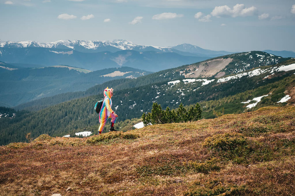 Полнометражный портрет туристки в костюме единорога, идущей через Карпатские горы, покрытые снежными остатками в солнечный весенний день. Концепция путешествий
. - Фото, изображение